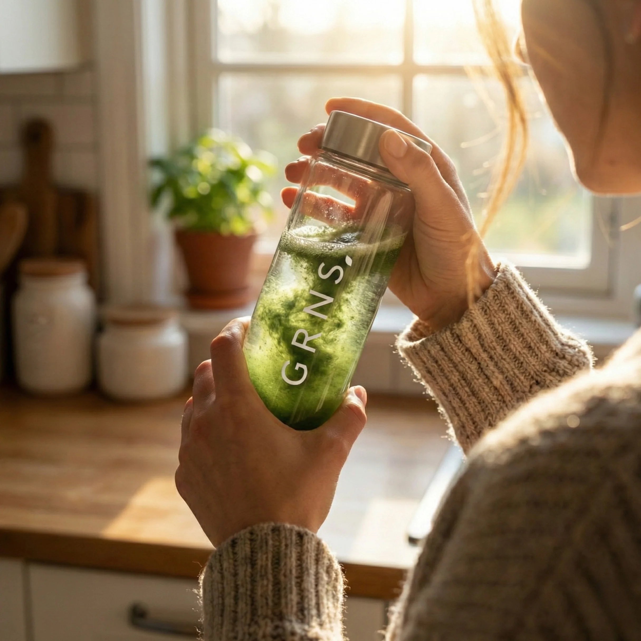 A smiling girl holding a glass of greens powder drink whilst having a green skincare mask on her face