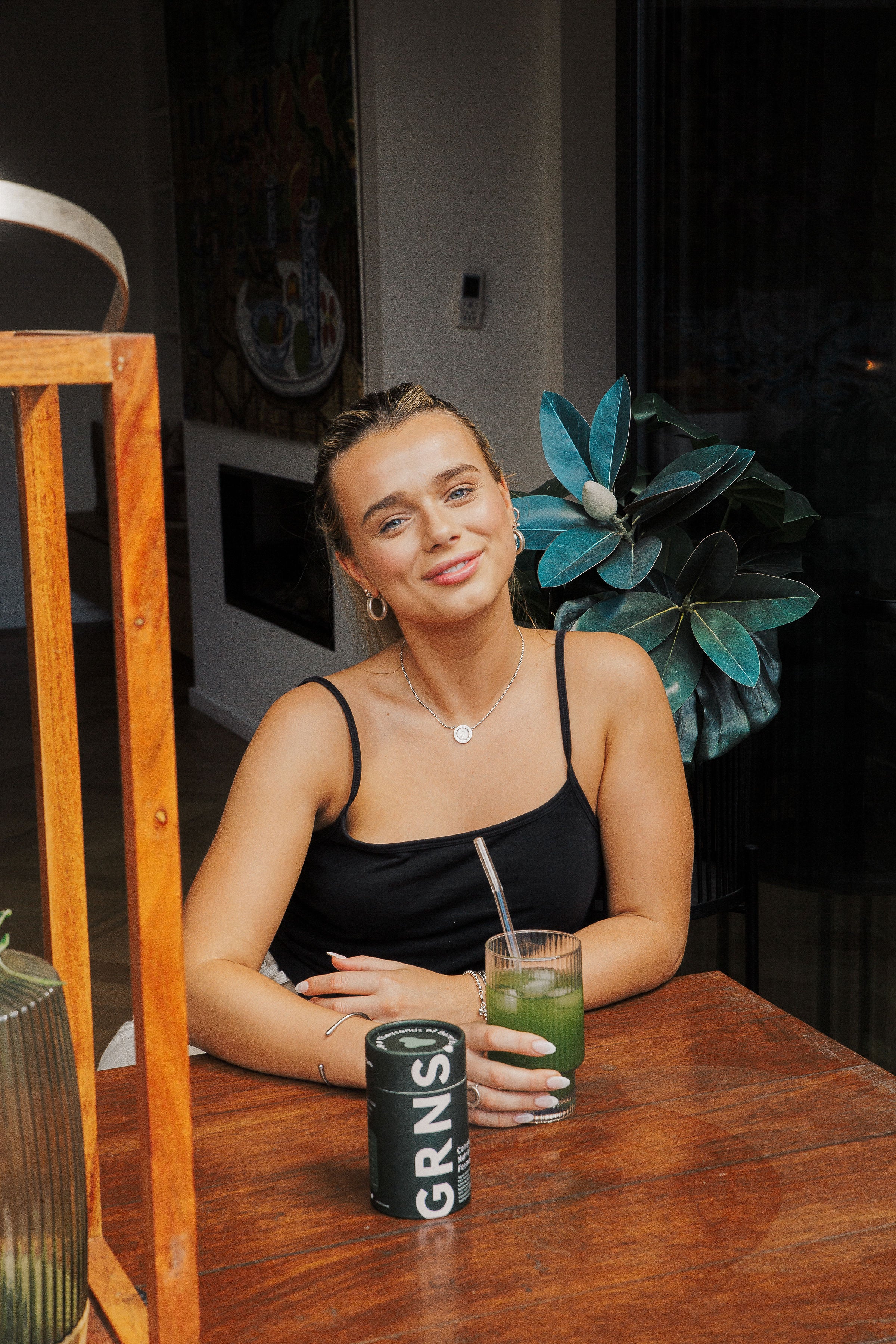 Smiling woman enjoying a refreshing GRNS green juice, seated indoors with vibrant greenery in the background, conveying health and relaxation