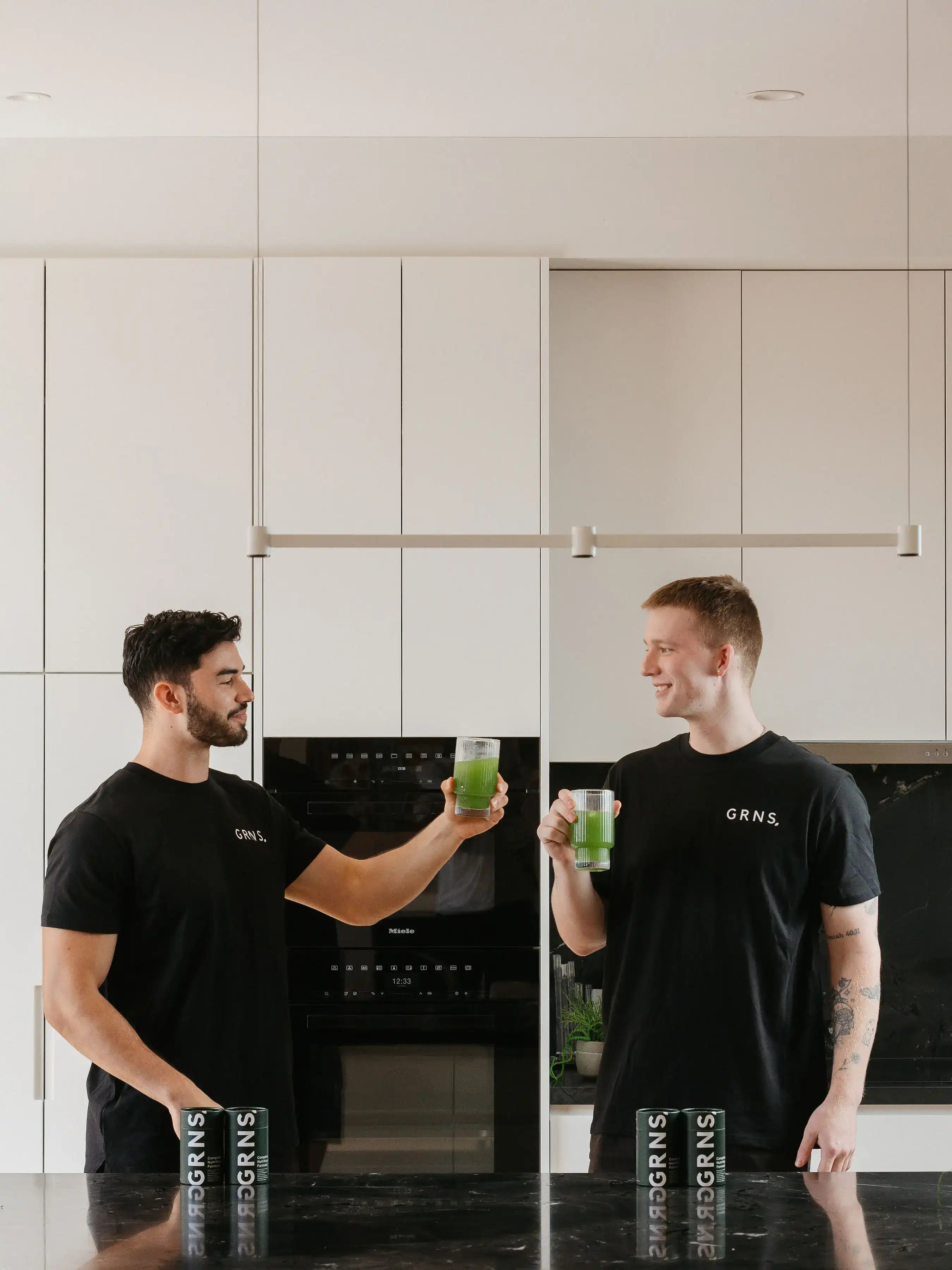 Two friends sharing a moment of connection, smiling and toasting with GRNS greens drinks in a modern kitchen setting, embodying health and happiness