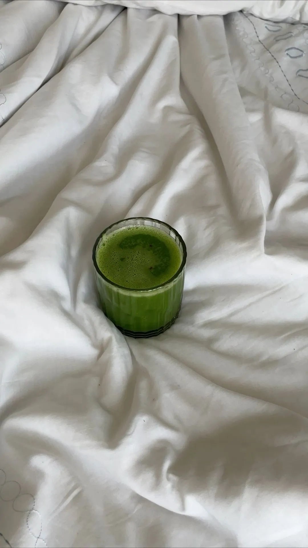 A glass of greens powder on a white bed