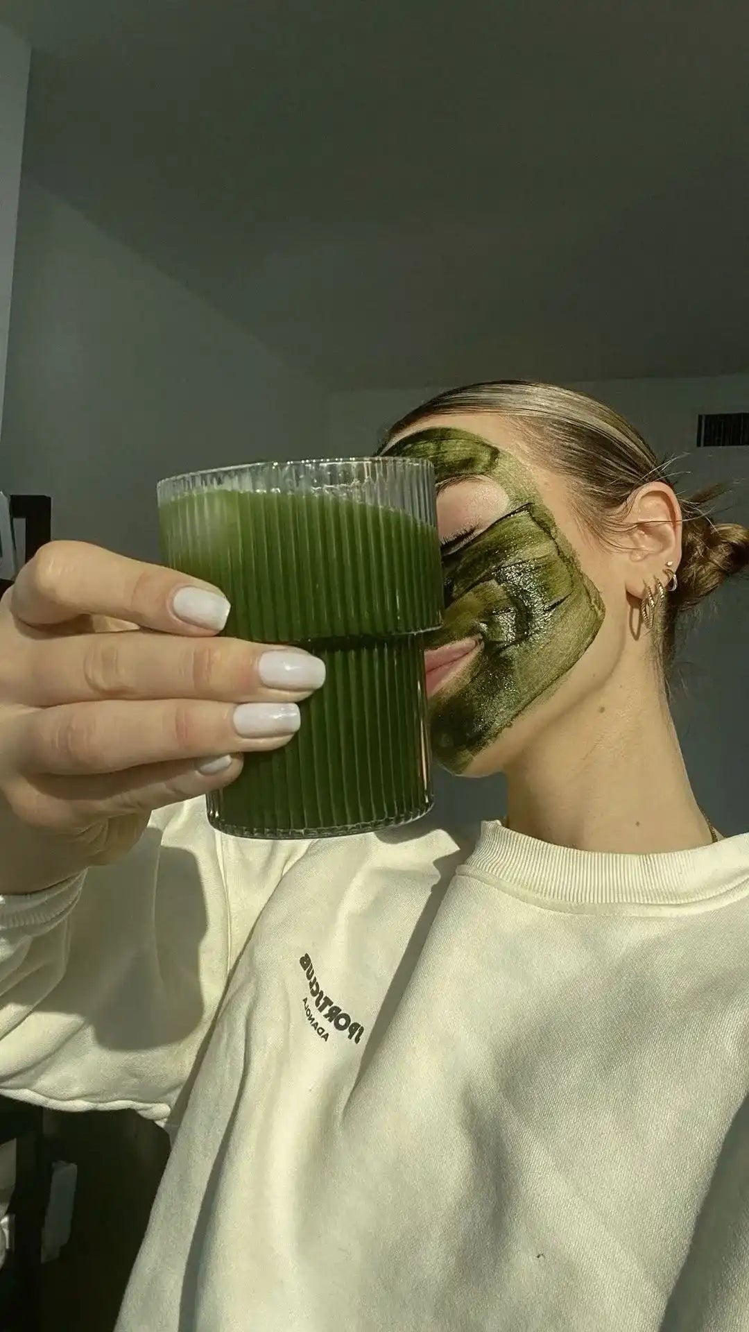 A smiling girl holding a glass of greens powder drink whilst having a green skincare mask on her face